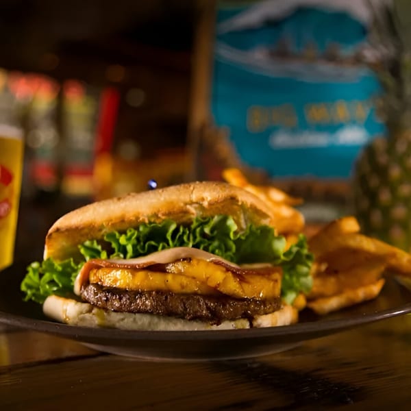 Wraptitude's burger with grilled pineapple, bacon, lettuce, and fries on a dark plate.