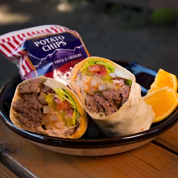 Two Wraptitude burger wraps, a bag of potato chips, and orange slices on a dark plate.