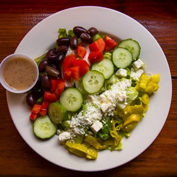 A fresh Greek salad with lettuce, tomatoes, cucumbers, olives, feta cheese, and pepperoncini, with dressing on the side.